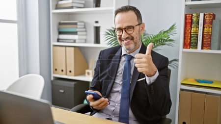 Photo for Cheerful, grey-haired middle age man, a diligent business worker, works his smartphone, giving an assertive thumb up gesture in his office room, projecting a confident, positive aura! - Royalty Free Image