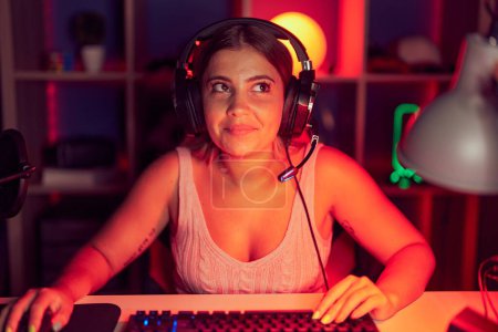 Téléchargez les photos : Jeune femme blonde jouant à des jeux vidéo portant un casque souriant regardant vers le côté et regardant loin en pensant. - en image libre de droit