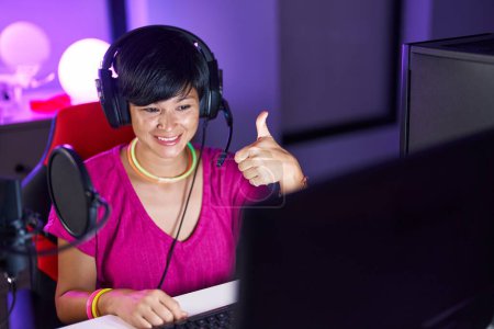 Foto de Joven mujer asiática con pelo corto jugando videojuegos sonriendo feliz y positivo, pulgar hacia arriba haciendo excelente y aprobación signo - Imagen libre de derechos