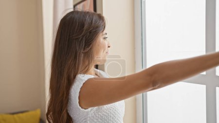 Photo for Joyful young hispanic woman, laughing and throwing glimpses outside, stands by the living room window at home with confident smile - Royalty Free Image