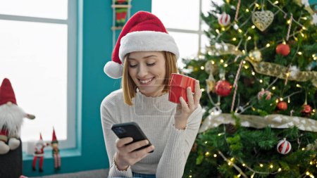 Foto de Mujer rubia joven usando smartphone bebiendo café celebrando la Navidad en casa - Imagen libre de derechos