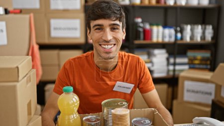 Foto de Retrato conmovedor de un joven y apuesto hombre hispano, sonriendo con confianza mientras se ofrece como voluntario en un centro de caridad, sentado en una mesa, lleno de donaciones en una vibrante sala comunitaria. - Imagen libre de derechos