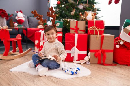 Téléchargez les photos : Adorable garçon hispanique écrivant santa claus lettre assis sur le sol par l'arbre de Noël à la maison - en image libre de droit