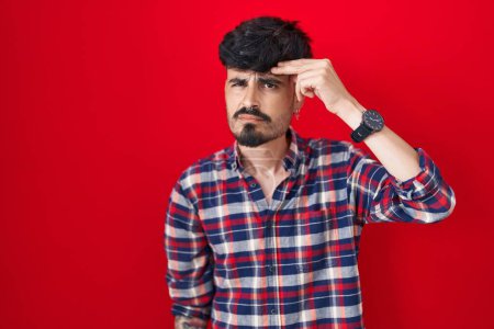 Photo for Young hispanic man with beard standing over red background pointing unhappy to pimple on forehead, ugly infection of blackhead. acne and skin problem - Royalty Free Image