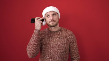 Photo for Young caucasian man listening to voice message wearing christmas hat over isolated red background - Royalty Free Image