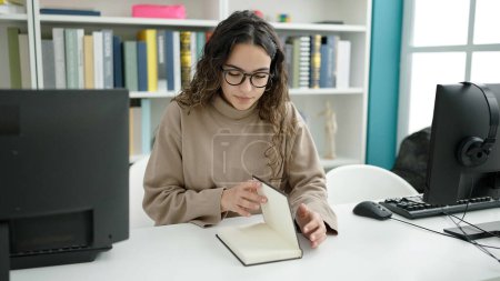 Téléchargez les photos : Jeune belle hispanique étudiant lecture livre à la bibliothèque université - en image libre de droit