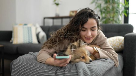 Photo for Young hispanic woman with dog using smartphone lying on sofa at home - Royalty Free Image