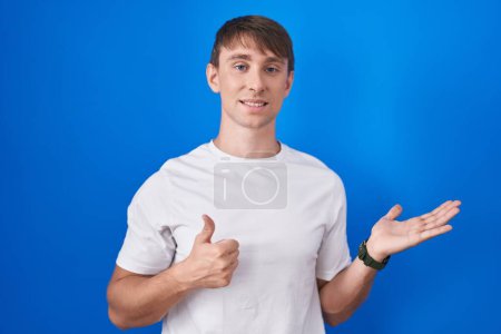 Téléchargez les photos : Homme blond caucasien debout sur fond bleu montrant la main de la paume et faisant un geste correct avec les pouces levés, souriant heureux et joyeux - en image libre de droit