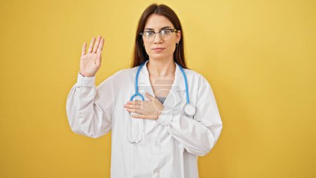 Téléchargez les photos : Jeune belle femme hispanique médecin faire un serment avec la main sur la poitrine sur fond jaune isolé - en image libre de droit