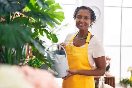 Téléchargez les photos : Moyen Age afro-américaine fleuriste souriant plante arrosante confiante à la boutique de fleurs - en image libre de droit