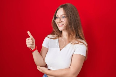Téléchargez les photos : Jeune femme caucasienne debout sur fond rouge regardant fier, souriant faire les pouces vers le haut geste sur le côté - en image libre de droit