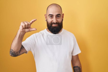 Téléchargez les photos : Jeune homme hispanique avec barbe et tatouages debout sur fond jaune geste souriant et confiant avec la main faisant signe de petite taille avec les doigts regardant et la caméra. concept de mesure. - en image libre de droit