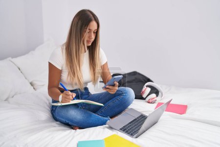 Téléchargez les photos : Jeune belle étudiante hispanique utilisant un smartphone écrivant sur un ordinateur portable dans la chambre - en image libre de droit