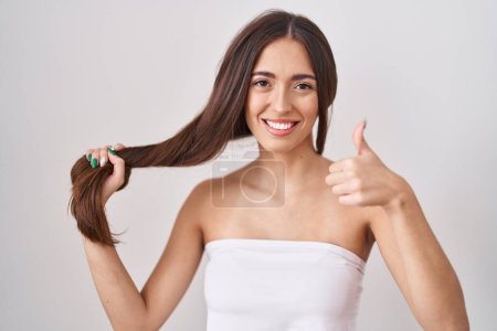 Téléchargez les photos : Jeune femme hispanique tenant les cheveux avec la main souriante heureuse et positive, pouce levé faire excellent et signe d'approbation - en image libre de droit