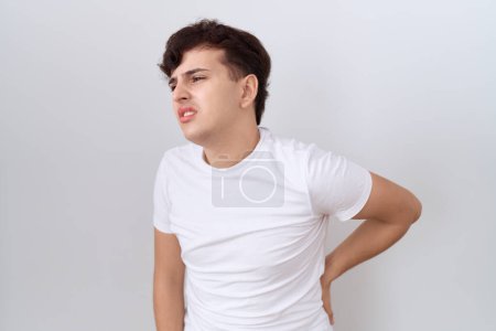 Téléchargez les photos : Jeune homme non binaire portant un t-shirt blanc décontracté souffrant de maux de dos, toucher le dos avec la main, douleur musculaire - en image libre de droit
