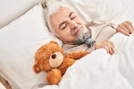 Photo for Middle age grey-haired man lying on bed sleeping with teddy beat at bedroom - Royalty Free Image