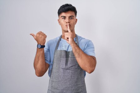 Photo for Hispanic young man wearing apron over white background asking to be quiet with finger on lips pointing with hand to the side. silence and secret concept. - Royalty Free Image