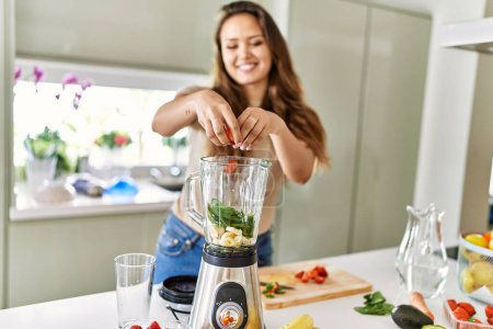 Photo for Young beautiful hispanic woman preparing vegetable smoothie with blender at the kitchen - Royalty Free Image