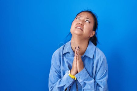 Téléchargez les photos : Asiatique jeune femme debout sur fond bleu mendier et prier avec les mains ainsi que l'expression de l'espoir sur le visage très émotionnel et inquiet. mendier. - en image libre de droit