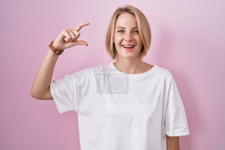 Foto de Joven mujer caucásica de pie sobre fondo rosa sonriente y seguro gesto con la mano haciendo signo de tamaño pequeño con los dedos mirando y la cámara. concepto de medida. - Imagen libre de derechos