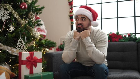 Téléchargez les photos : Jeune homme hispanique assis sur le canapé près de l'arbre de Noël avec une expression triste à la maison - en image libre de droit