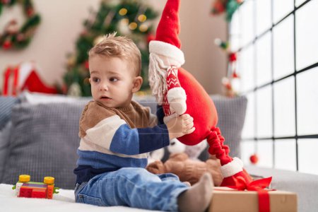 Photo for Adorable toddler playing with santa claus doll sitting on sofa by christmas tree at home - Royalty Free Image