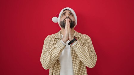 Photo for Young hispanic man praying wearing christmas hat over isolated red background - Royalty Free Image