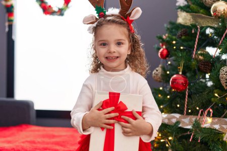 Foto de Adorable chica rubia abrazando regalo de pie junto al árbol de Navidad en casa - Imagen libre de derechos