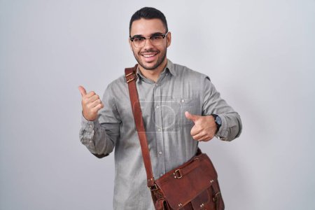 Photo for Young hispanic man wearing suitcase pointing to the back behind with hand and thumbs up, smiling confident - Royalty Free Image