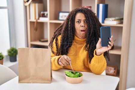 Photo for Plus size hispanic woman eating take away salad holding smartphone afraid and shocked with surprise and amazed expression, fear and excited face. - Royalty Free Image