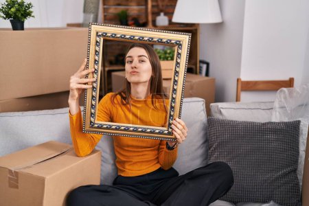 Photo for Young caucasian woman at new home holding empty frame looking at the camera blowing a kiss being lovely and sexy. love expression. - Royalty Free Image