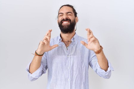 Téléchargez les photos : Homme hispanique à la barbe portant une chemise décontractée geste doigt croisé souriant avec espoir et les yeux fermés. chance et concept superstitieux. - en image libre de droit