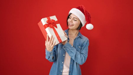 Foto de Joven mujer caucásica sonriendo confiada usando sombrero de Navidad sobre fondo rojo aislado - Imagen libre de derechos