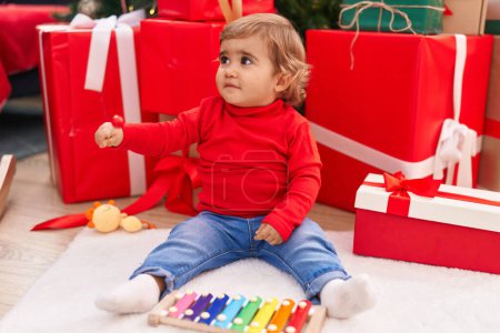 Photo for Adorable hispanic toddler playing xylophone sitting on floor by christmas gifts at home - Royalty Free Image