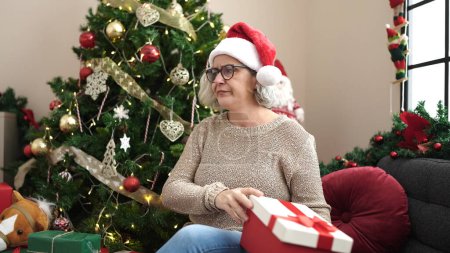 Téléchargez les photos : Femme d'âge moyen avec les cheveux gris portant le chapeau de Noël déballer cadeau regardant bouleversé à la maison - en image libre de droit