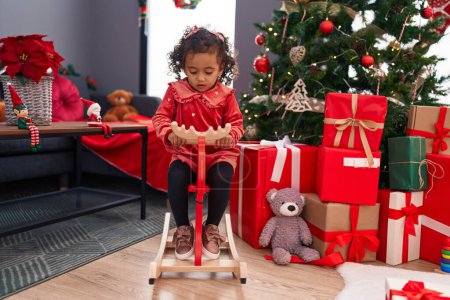 Photo for Adorable hispanic girl playing with reindeer rocking by christmas tree at home - Royalty Free Image