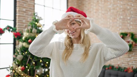 Photo for Young blonde woman celebrating christmas doing heart shape with hands at home - Royalty Free Image
