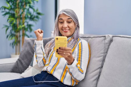 Photo for Middle age woman with grey hair using smartphone sitting on the sofa screaming proud, celebrating victory and success very excited with raised arm - Royalty Free Image
