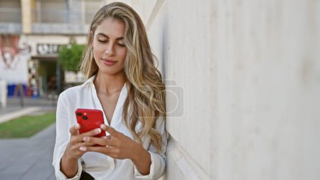 Téléchargez les photos : Chic jeune femme blonde avec une expression sérieuse absorbée dans son smartphone, debout sur un fond de rue de la ville ensoleillée, illustrant le style de vie urbain cool et technicien. - en image libre de droit