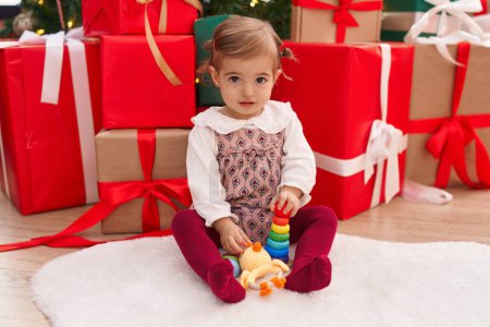 Téléchargez les photos : Adorable blond tout-petit sourire confiant assis sur le sol par des cadeaux de Noël à la maison - en image libre de droit