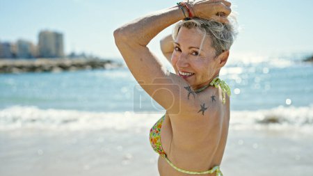 Foto de Mujer rubia de mediana edad turista sonriendo confiado tocando el pelo en la playa - Imagen libre de derechos