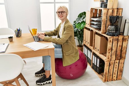 Foto de Joven mujer rubia trabajadora de negocios utilizando portátil sentado en la bola en forma en la oficina - Imagen libre de derechos