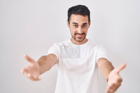 Foto de Hombre hispano guapo de pie sobre fondo blanco sonriente alegre ofreciendo manos dando asistencia y aceptación. - Imagen libre de derechos