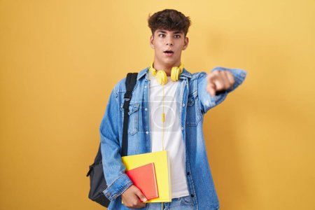 Téléchargez les photos : Adolescent hispanique portant un sac à dos étudiant et tenant des livres pointant du doigt surpris devant, bouche ouverte expression étonnée, quelque chose sur le devant - en image libre de droit