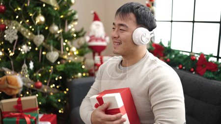 Téléchargez les photos : Jeune homme chinois écoutant de la musique tenant cadeau par arbre de Noël à la maison - en image libre de droit