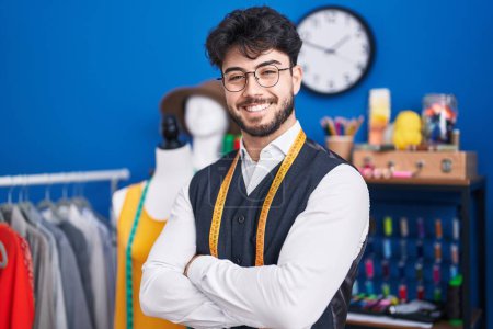 Foto de Joven hombre hispano sastre sonriendo confiado de pie con los brazos cruzados gesto en el estudio de costura - Imagen libre de derechos