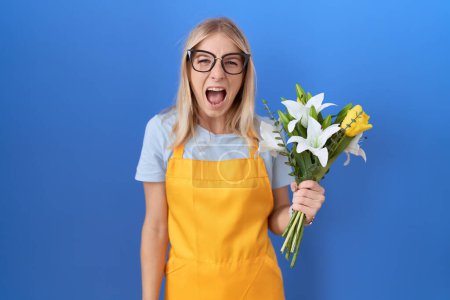 Foto de Young caucasian woman wearing florist apron holding flowers angry and mad screaming frustrated and furious, shouting with anger. rage and aggressive concept. - Imagen libre de derechos