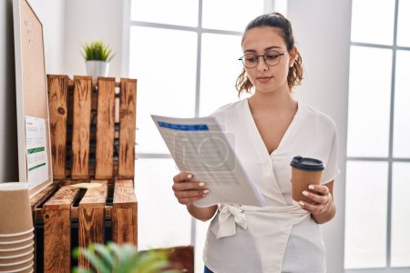 Foto de Joven hermosa mujer hispana trabajadora de negocios leyendo documento bebiendo café en la oficina - Imagen libre de derechos