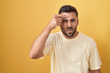Photo for Handsome hispanic man standing over yellow background pointing unhappy to pimple on forehead, ugly infection of blackhead. acne and skin problem - Royalty Free Image