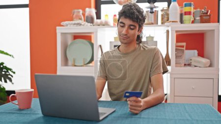 Foto de Joven hombre hispano de compras con portátil y tarjeta de crédito sentado en la mesa en el comedor - Imagen libre de derechos
